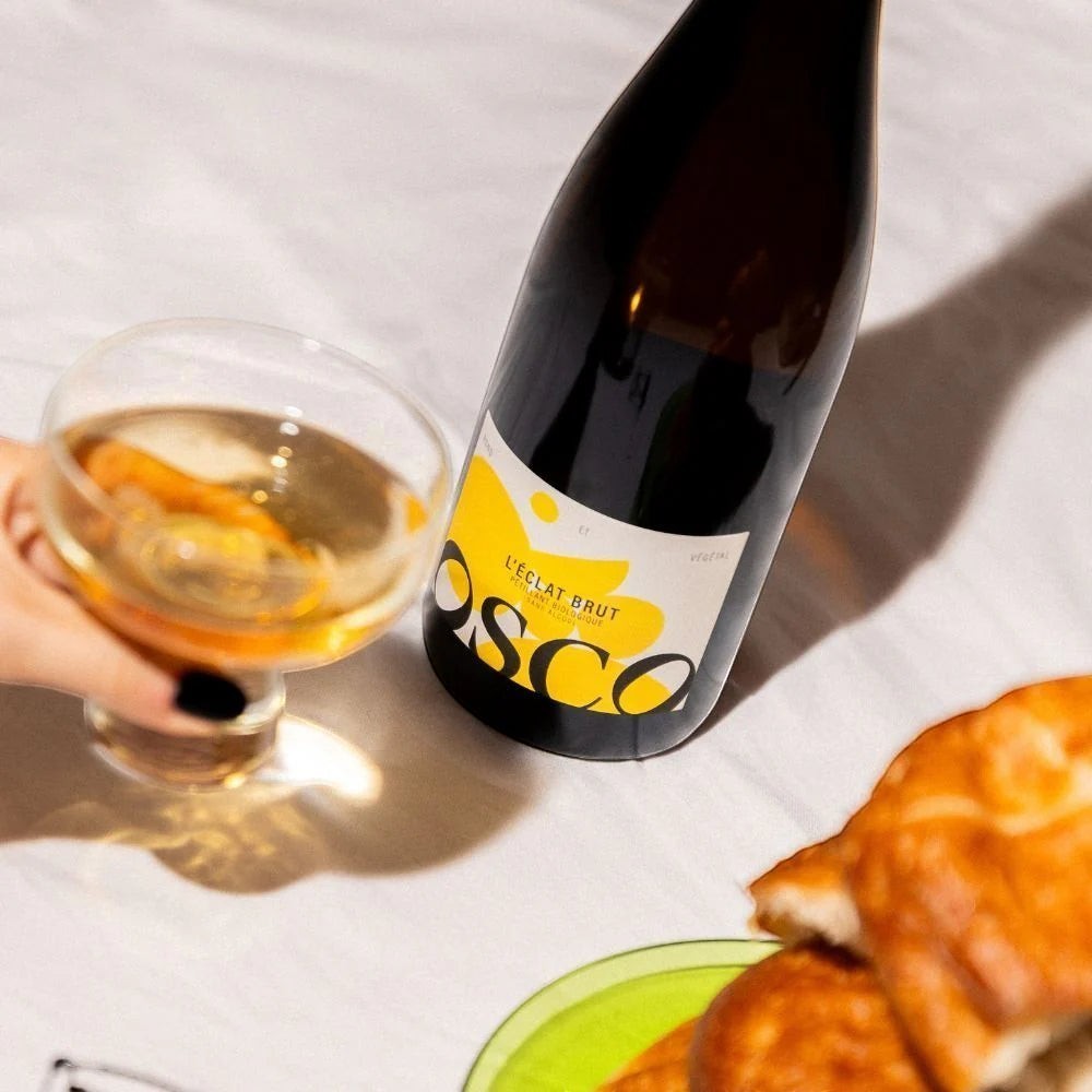 OSCO L'Éclat Brut non-alcoholic sparkling drink bottle next to a glass and pastries on a white tablecloth